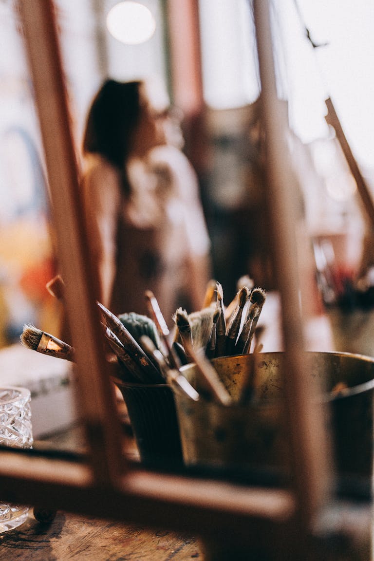 Blurred art studio scene with paintbrushes and background figure, evoking creativity and focus.
